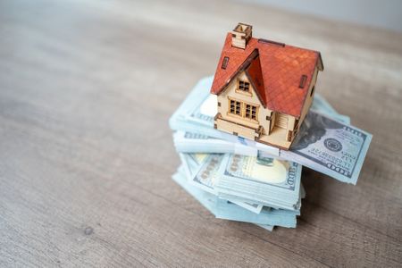 miniature house on stack of bills on wooden surface