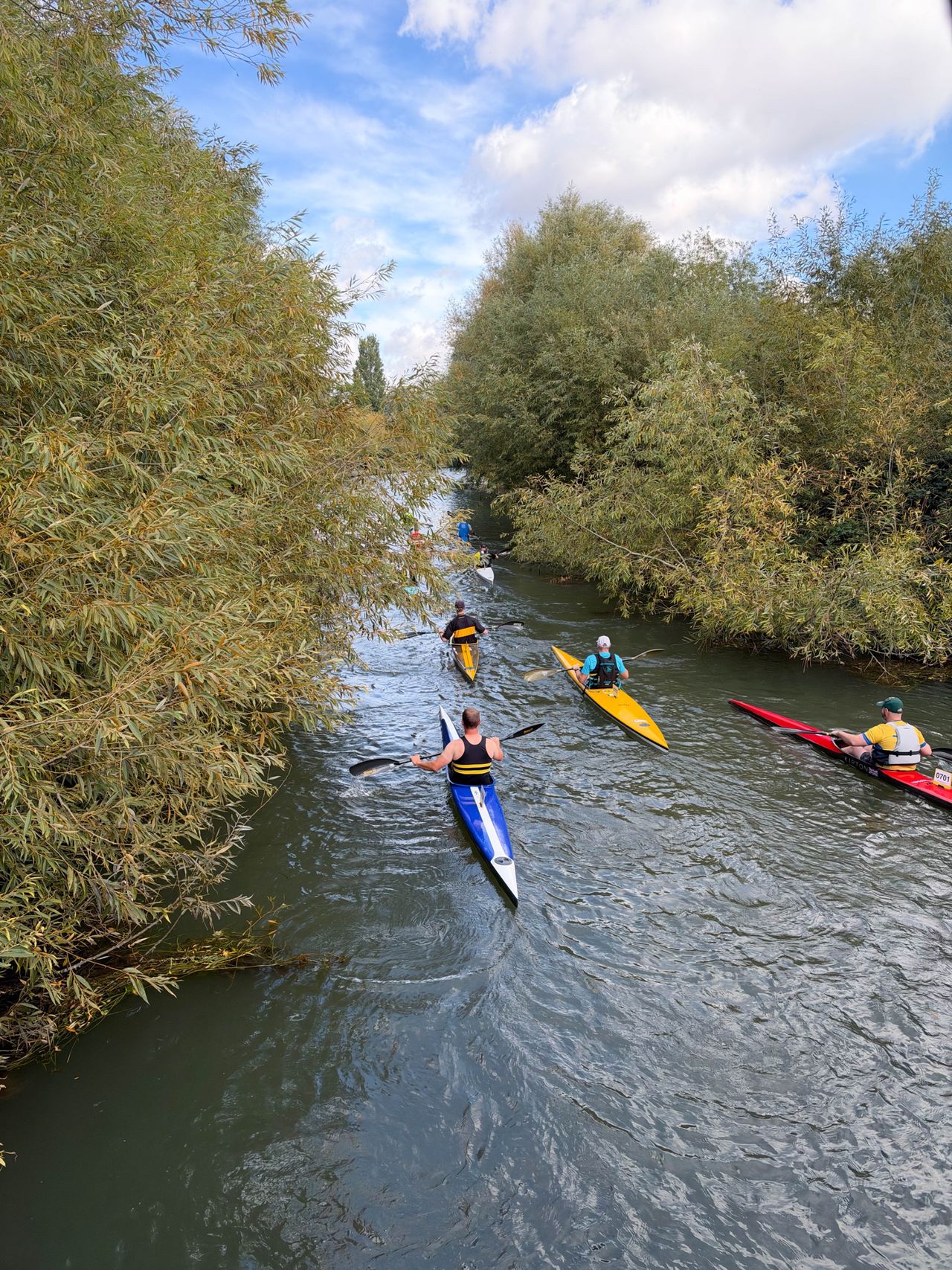 Several kayakers paddling along a narrow river bordered by dense willow trees beneath a partly cloudy sky.