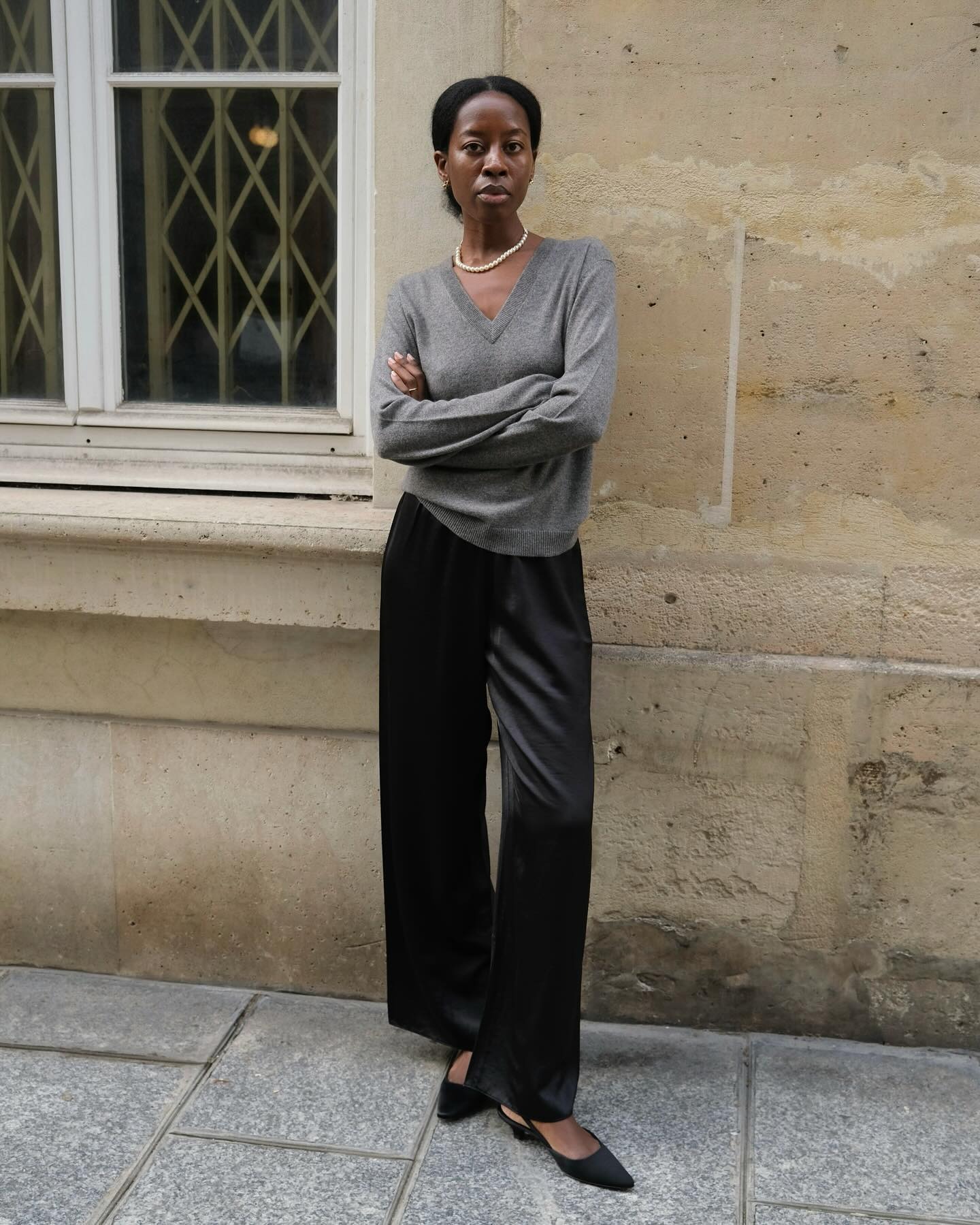 Style influencer Sylvie Mus poses on a Paris sidewalk wearing a pearl necklace, a gray v-neck sweater, relaxed black satin pants, and black kitten-heel slingback heels.