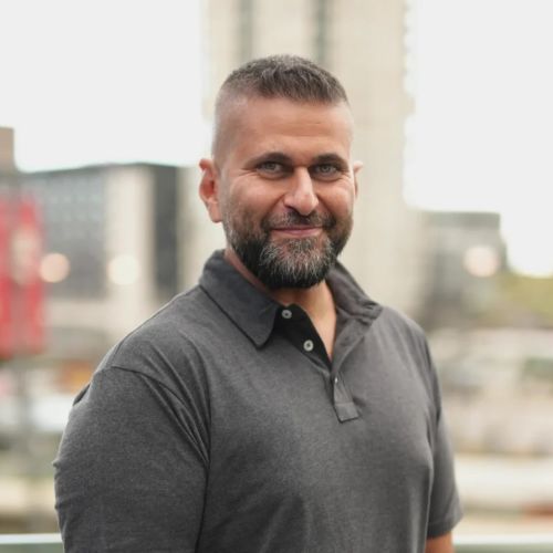 Headshot of Michael Zohouri wearing gray polo shirt, stood in front of blurred-out city background.