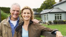 An older couple poses in front of their rural home.