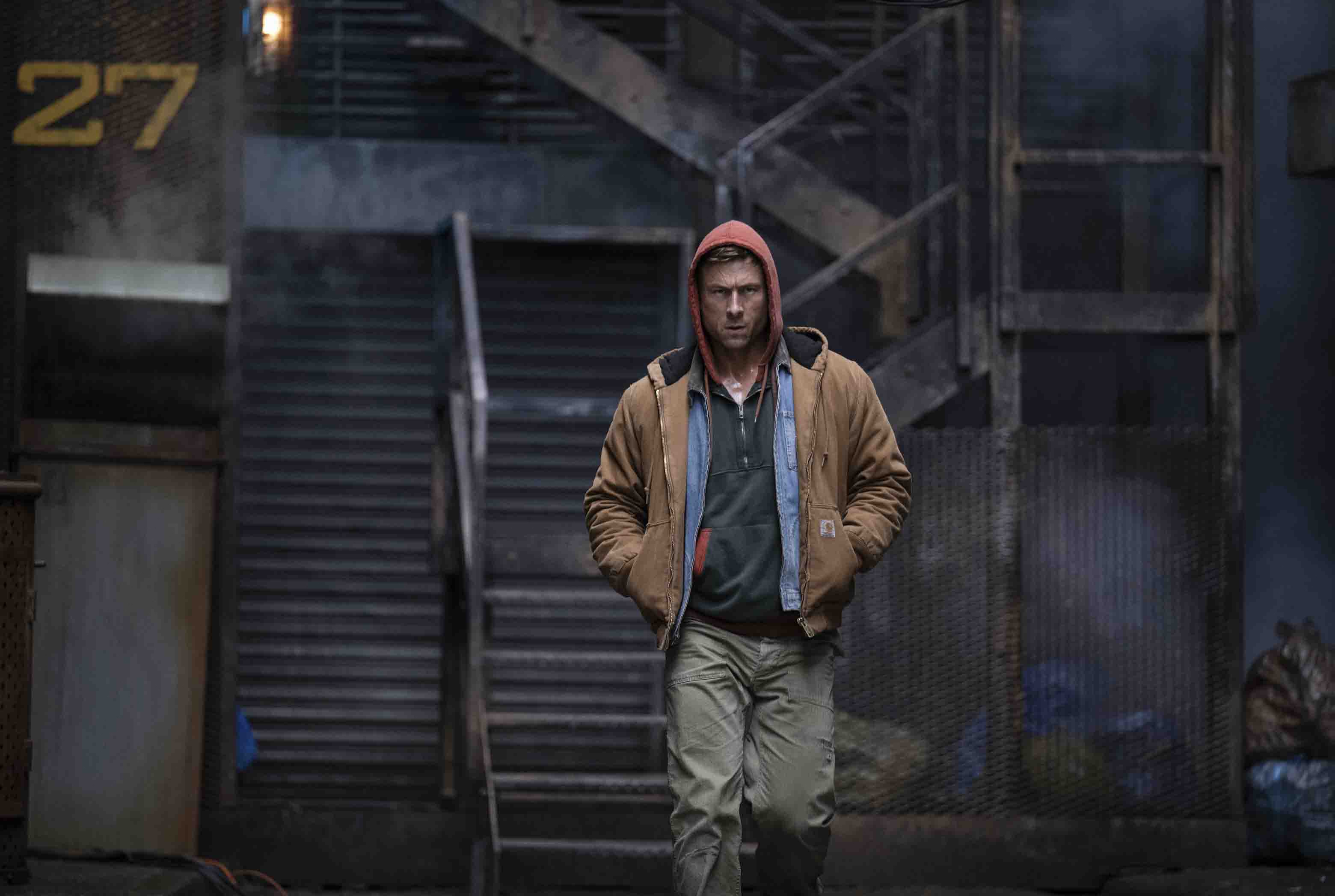 glen powell wearing a carhart jacket and hoodie walking down a dark alley in a still from the movie the running man