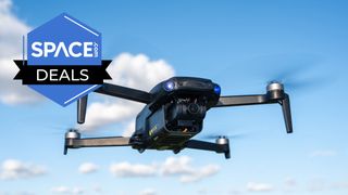 A black quadcopter is mid-flight and hovering in front of a blue sky background that features a handful of small clouds. The space.com logo is in the top left of the picture.