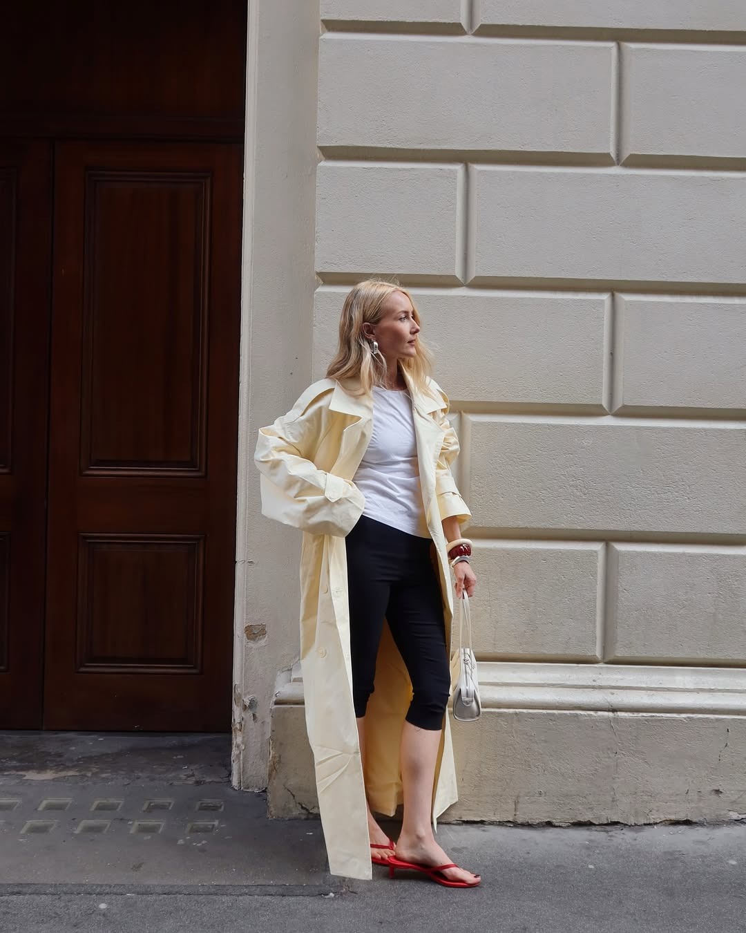 La mujer lleva una gabardina amarilla, una camiseta blanca, unos pantalones capri negros y unas sandalias rojas.