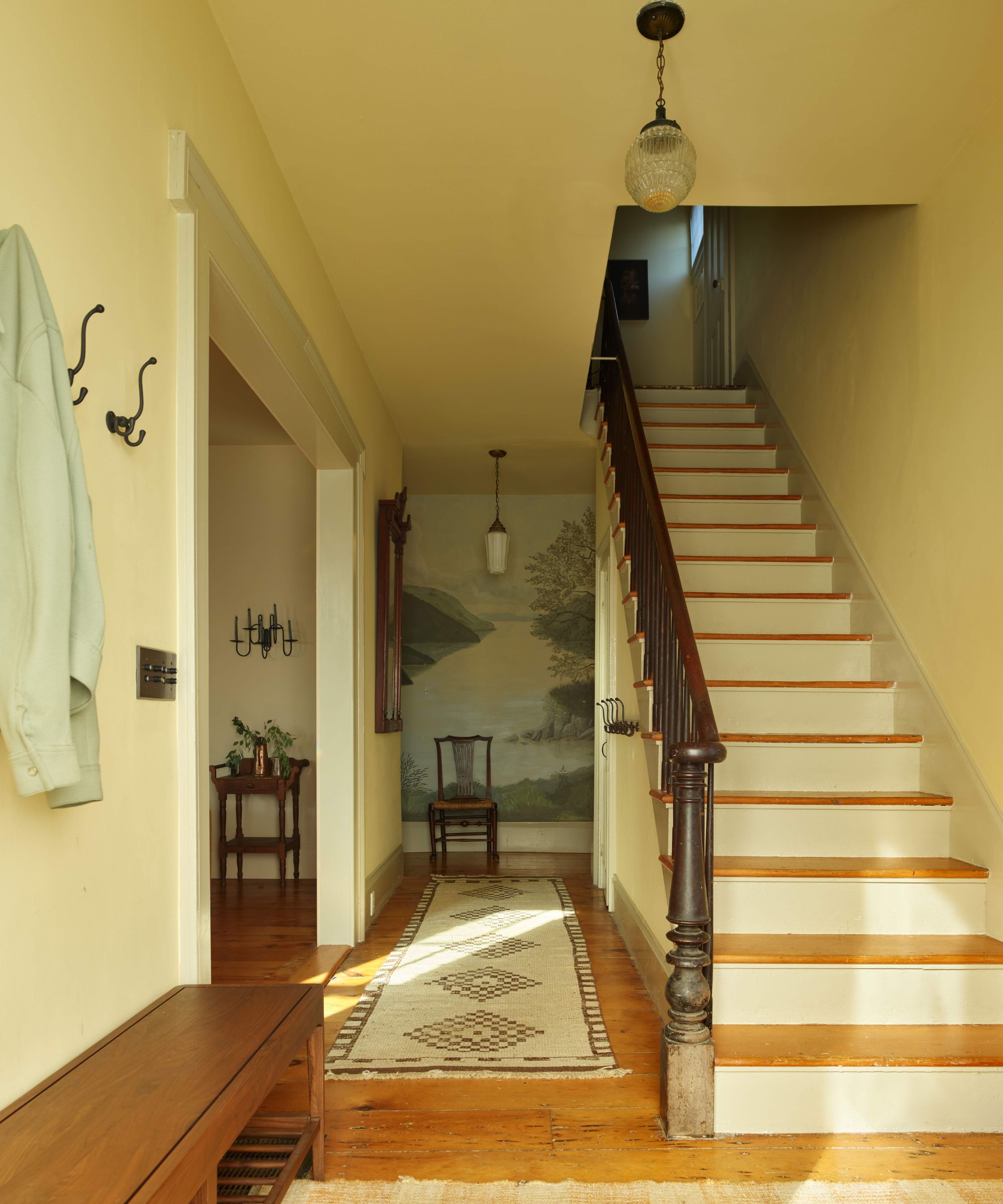 Sunlit hallway/foyer with pale yellow walls, a wooden staircase with dark railing, a landscape mural on the wall, and a low wooden bench
