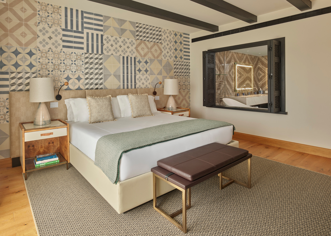 A bedroom with a colorful backdrop tiling behind the bed. There are also bedside tables with lamps on top, as well as a bench by the foot of the bed.