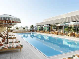 View of the rooftop pool at White City House. There are multicoloured floral lounge chairs with parasols on one side of the pool and tables and chairs with parasols on the other side under a clear blue sky