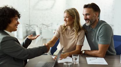 An insurance agent smiles as she shakes the hand of the woman while meeting a couple in her office.