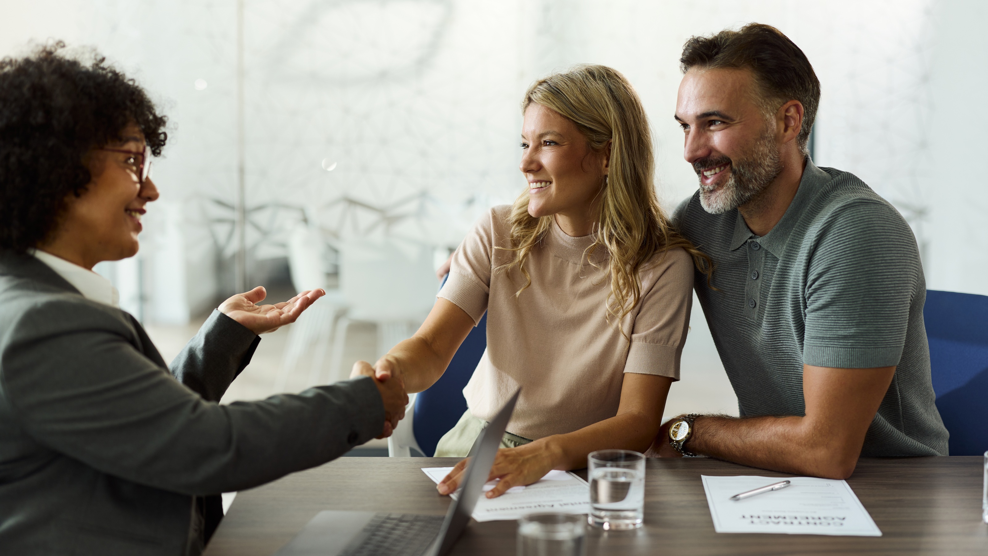 An insurance agent smiles as she shakes the hand of the woman while meeting a couple in her office.