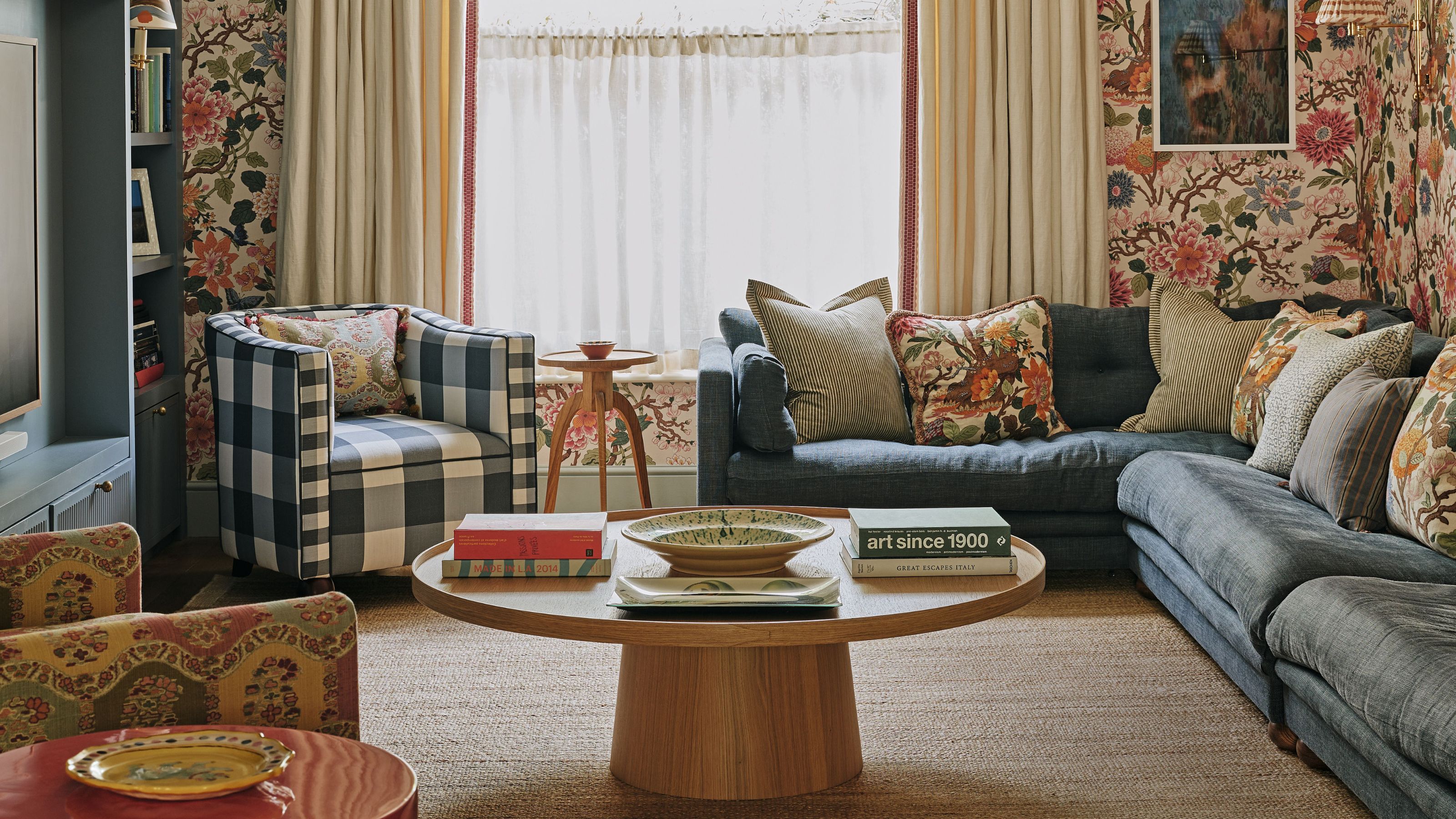 A cozy, maximalist living room featuring floral wallpaper, a blue l-shaped sofa with mismatched pillows, and a large circular oak coffee table on a jute rug.