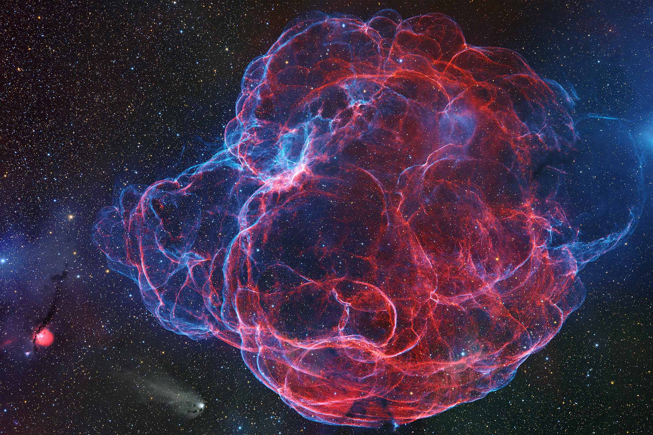 A colorful nebula blooms in space, displaying vibrant red and blue gases among countless stars against a dark cosmic backdrop