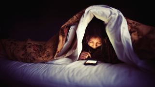 A young woman under the duvet in bed, staring at her phone.