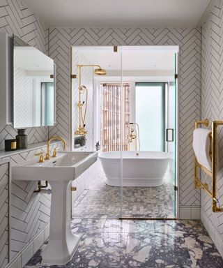 Large bathroom with separate shower and bath area, divided by glass doors. White herringbone patterned tiles on walls, marble flooring, brass fixtures and fittings.