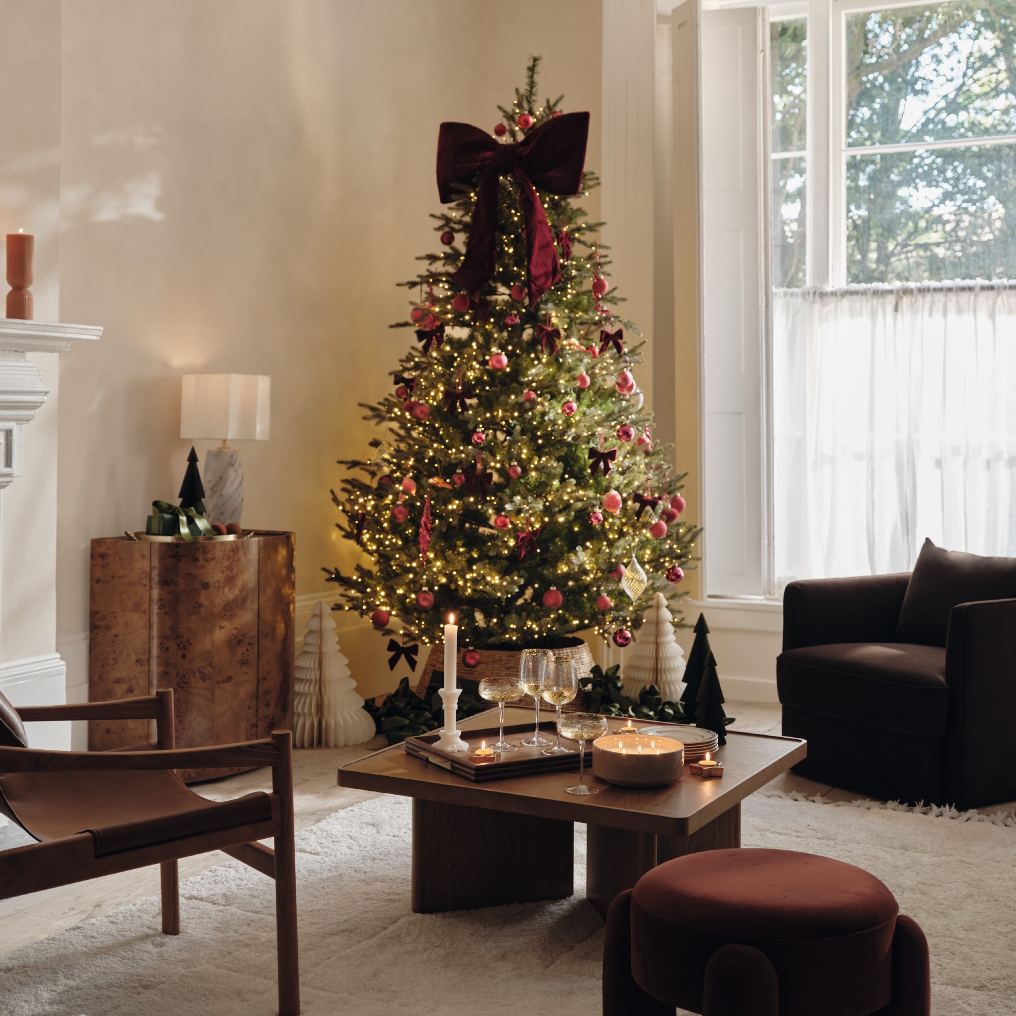 A modern living room decorated for Christmas with a lit-up Christmas tree in the corner decorated with a large red velvet bow