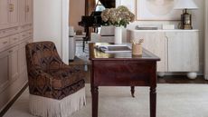 home office with beige carpet, built in cabinetry painted pink with a dark wood antique desk and small upholstered armchair with fringe trim