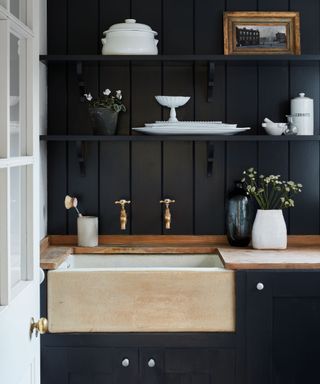 Dark laundry room ideas with black shiplap walls, white accessories, open shelving and a natural wood-clad sink.
