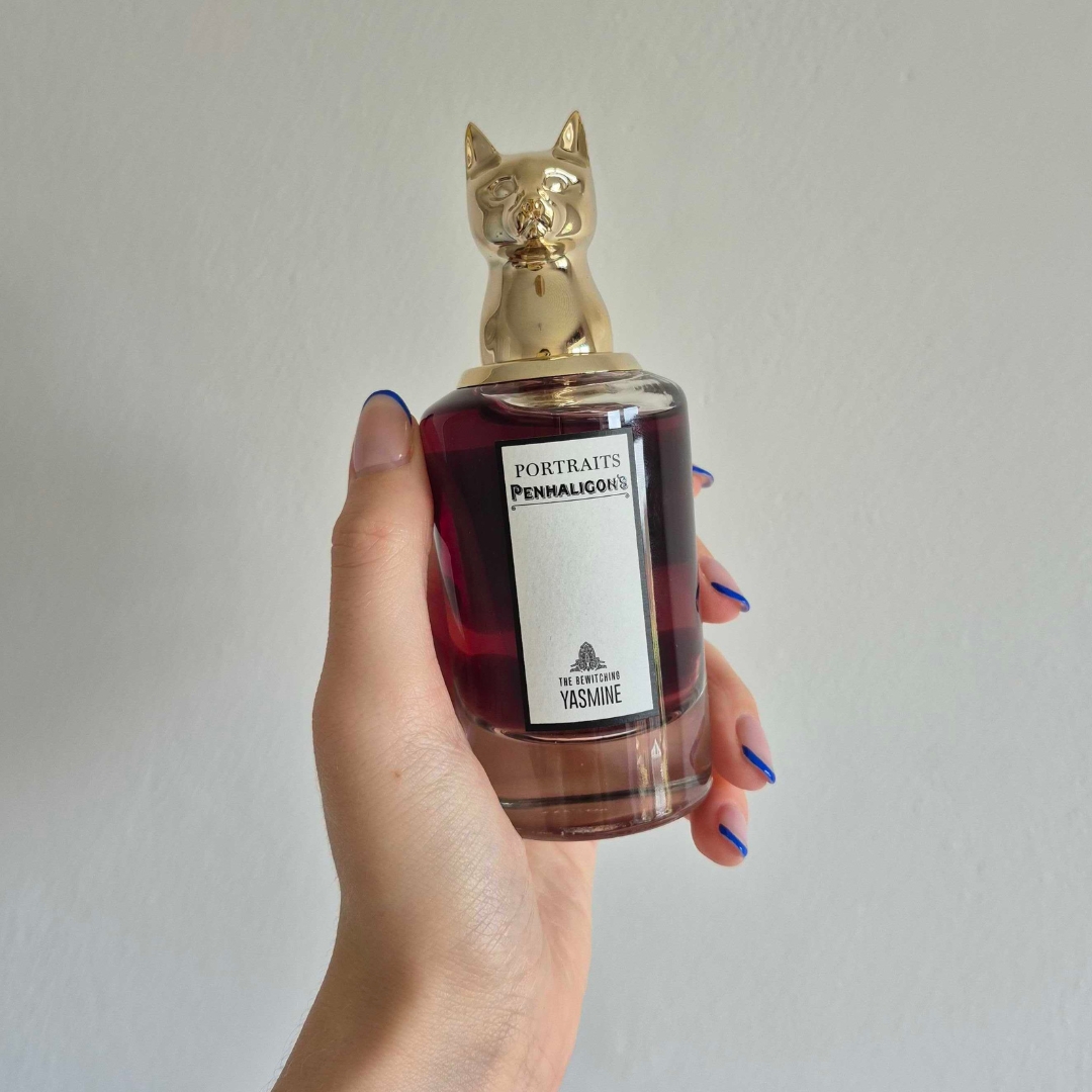 A close-up of Digital Beauty Writer Naomi Jamieson's hand, seen holding a bottle of Penhaligon's The Bewitching Jasmine perfume, which features a gold cat's head sculpture lid/ in front of a cream wall background
