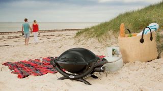 A Weber Baby Q barbecue is placed on the beach among a picnic.