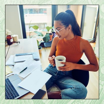 A young woman holds a cup of coffee while sitting at a desk and going through her taxes.