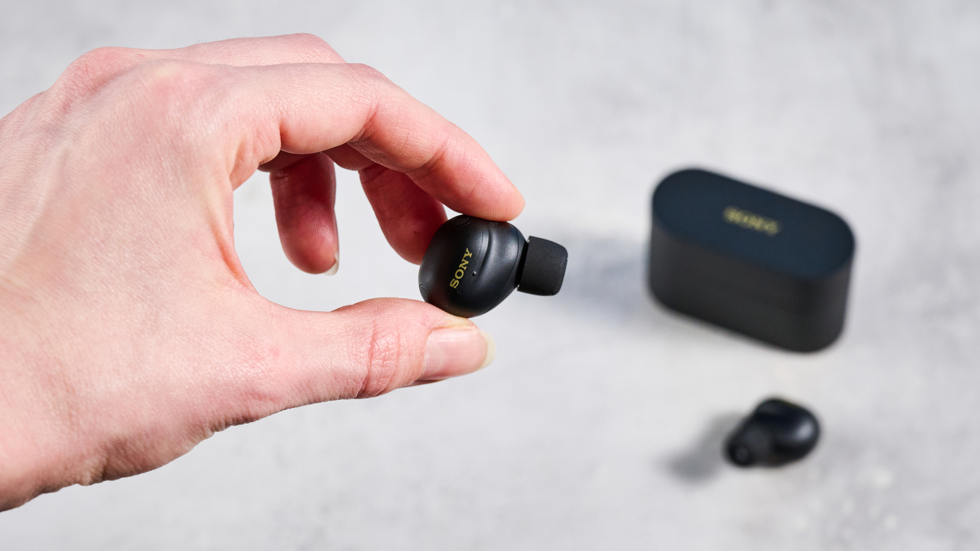 A hand holds a Sony WF-1000XM6 earbud, with the case and remaining bud out of focus in the background.