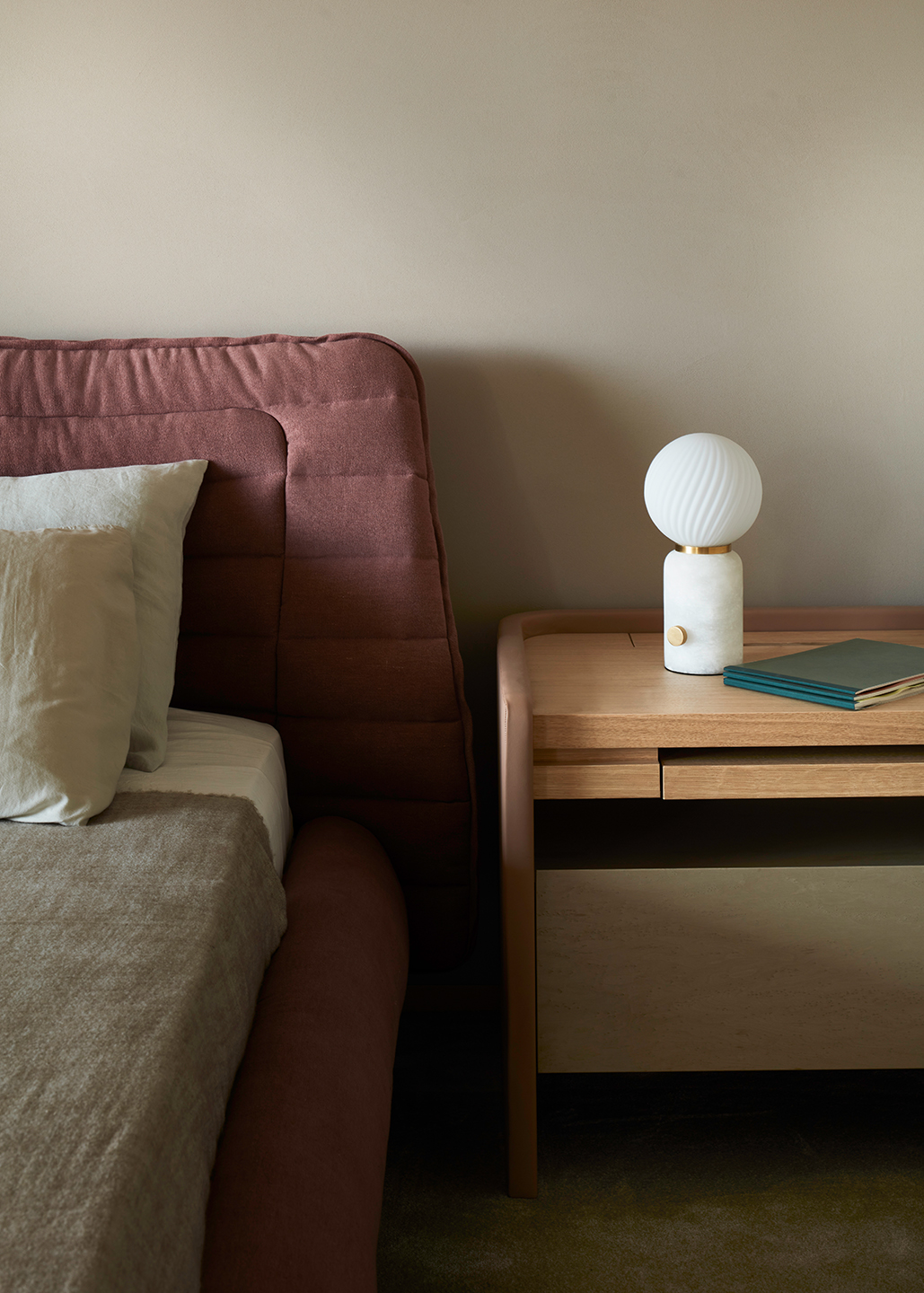 A close-up of a bordeaux, textured headboard with sage green sheets and a wooden bed-side table covered in ocean green notebooks and a white table lamp with a spherical top.
