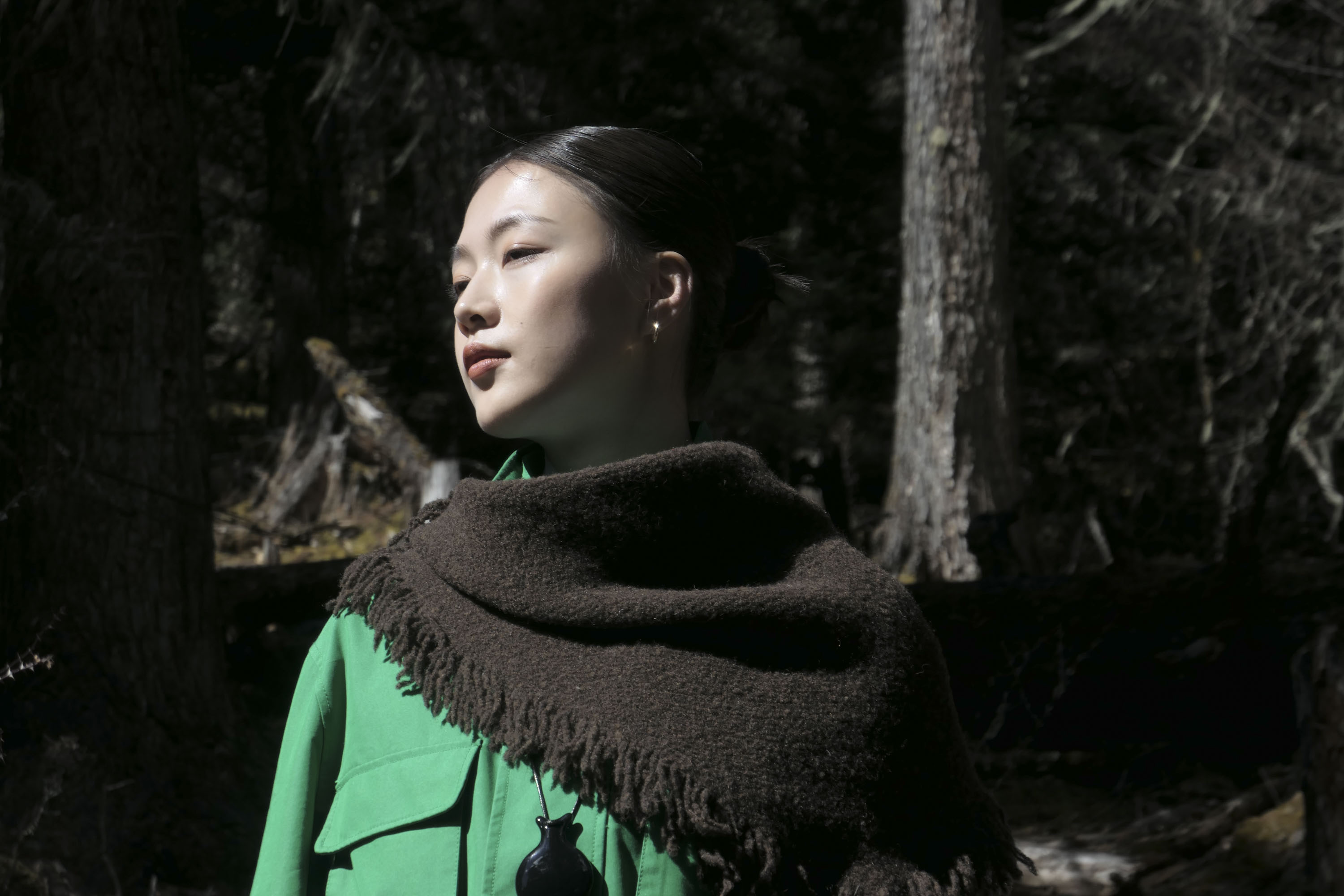 Portrait of a woman in a shawl and green jacket in a woodland, there's direct sunlight on her