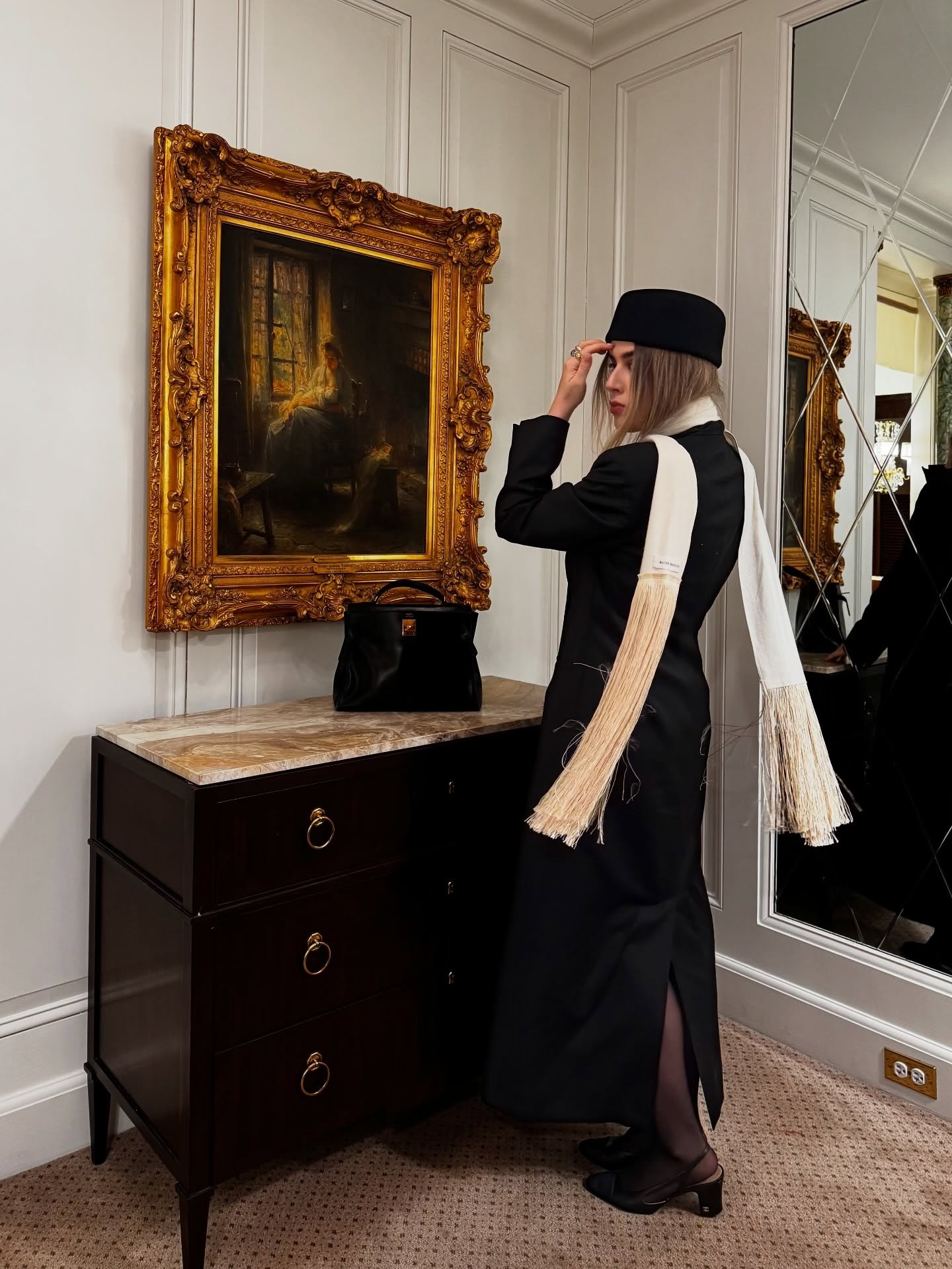 Woman wearing black coat, black pillbox hat, and white cream fringe scarf.