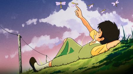a young boy lays on a grass hill reaching up at dragonflies
