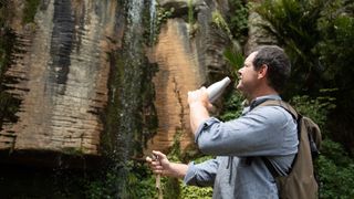 A hiker drinks water as he looks at a waterfall