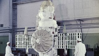 two scientists in white lab coats stand next to a cylindrical spacecraft in a hangar