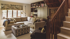 a living room with a large cream-colored couch, a square ottoman, a wall of bookshelves, a dark brown accent chair, a set of four windows in the background, and a wooden staircase in the foreground