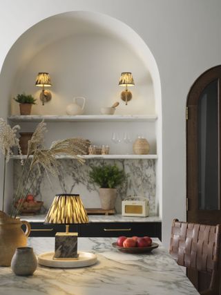 A kitchen with alcove shelving with two sconces with pleated shades, and a similar table lamp on the island