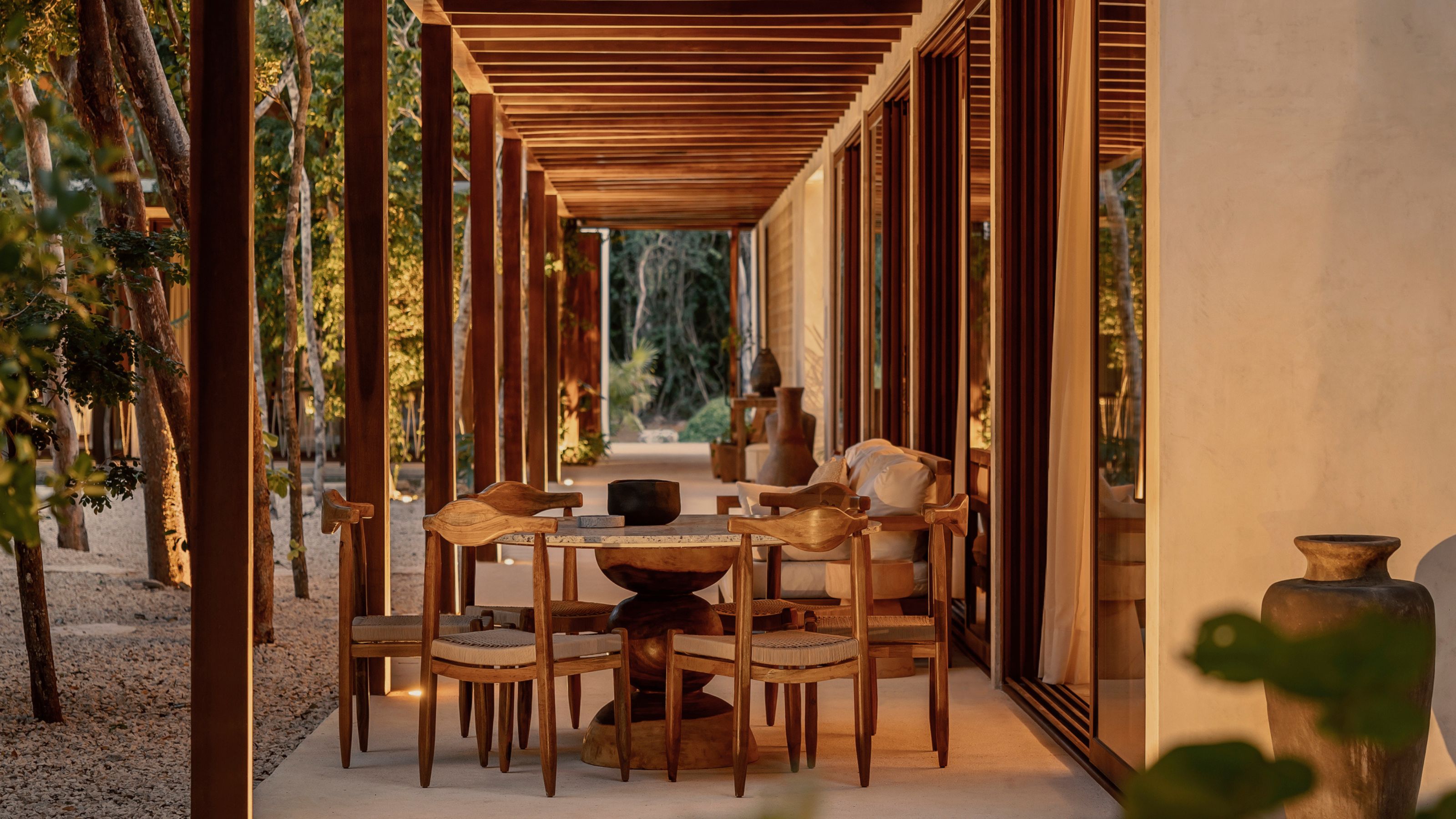 A covered outdoor dining area lit up with magical lighting at dusk with trees running along the lefthand side and a large pot just out of view on the opposite corner