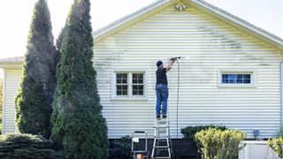 Man cleaning the side of his house with a pressure washer on a step ladder