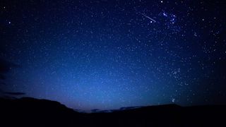A star-studded sky glitters above an undulating, dark, silhouetted horizon. The sky is a lighter shade of blue towards the horizon, where the outlines of clouds can be seen hugging the skyline. A lone meteor is visible streaking Earthwards towards the upper left of the image.
