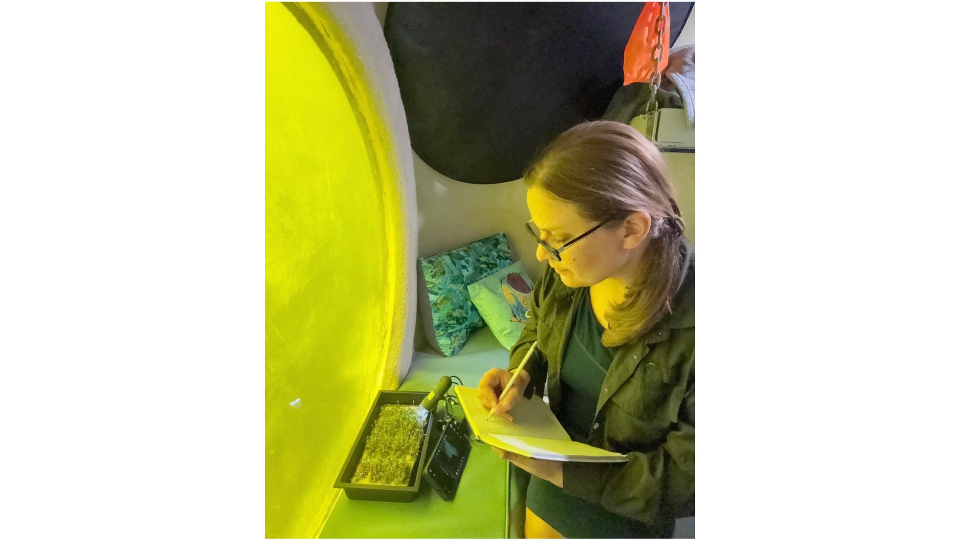 a woman makes notes about the growth of plants inside an underwater research station, next to a circular window looking out into greenish water