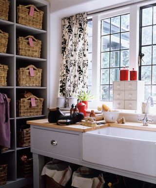 A utility room with a large farmhouse sink, grey cabinets and shelving filled with woven baskets