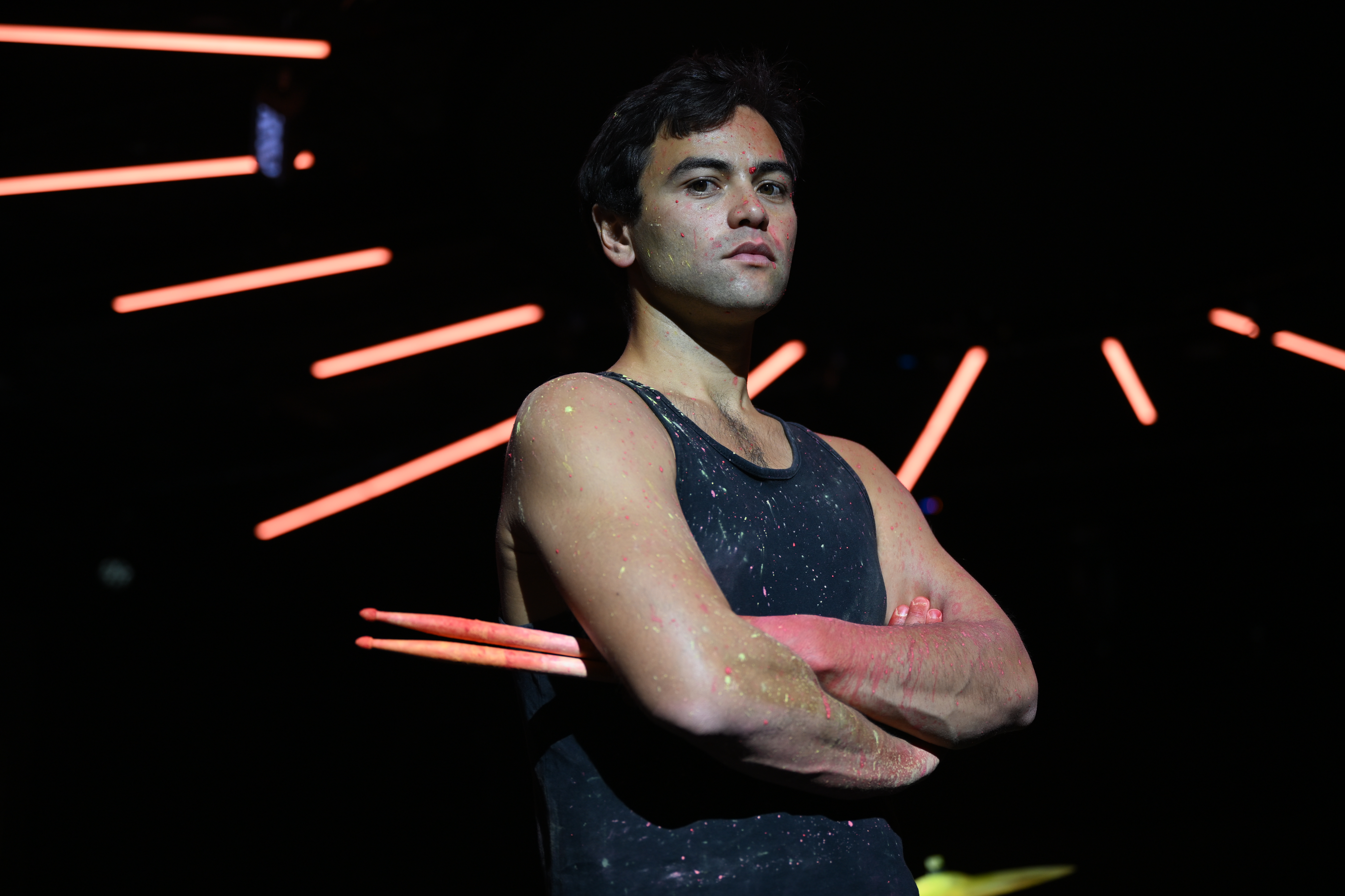 A male drummer in a dark studio, holding drums sticks with arms folded, neon lights in background