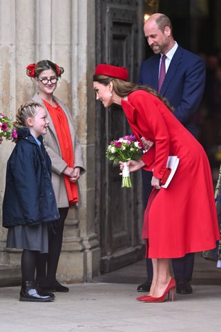 Kate Middleton wearing a red dress coat and hat bending forward to accept flowers from a little girl at Westminster Abbey in March 2025