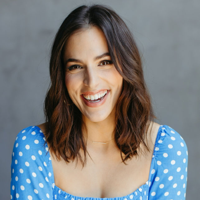 Woman with short brown hair smiling wearing blue and white polka dot top on a gray background is Janelle Cohen