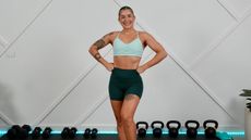 Trainer Roxanne Russell stands in front of rows of kettlebells and dumbbells in a studio, smiling.