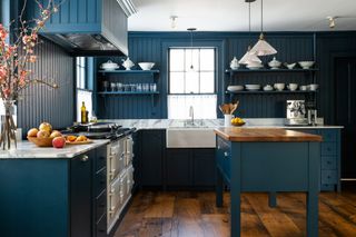 A dark teal blue kitchen with wall panelling, island and cabinetry all painted the same shade, a wooden floor and white worktops and Aga.