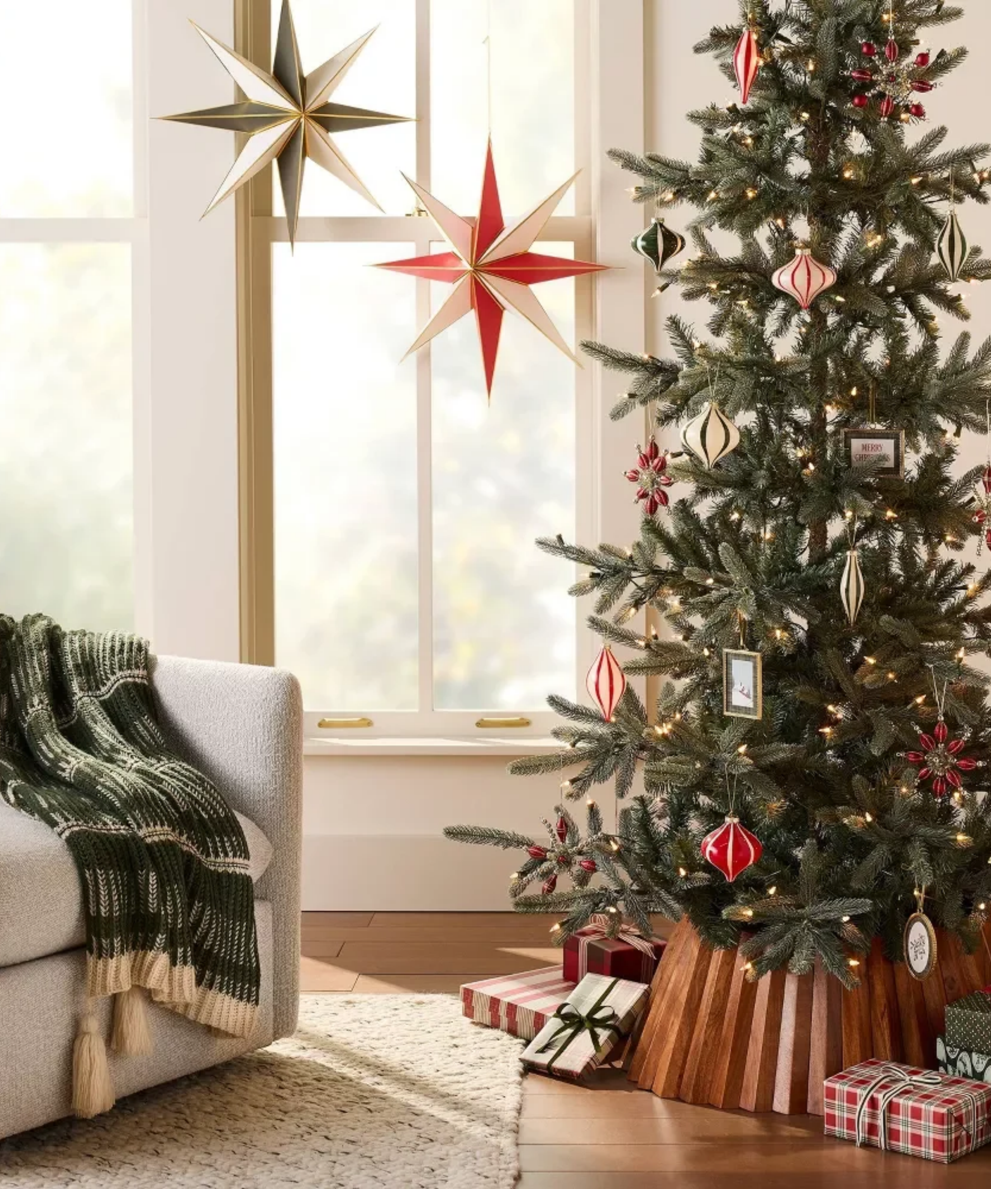 Retro-style holiday living room vignette, featuring a tree styled with a wooden tree skirt, hand-painted striped ornaments, an accent chair draped with a green knit throw, and paper lanterns overhead
