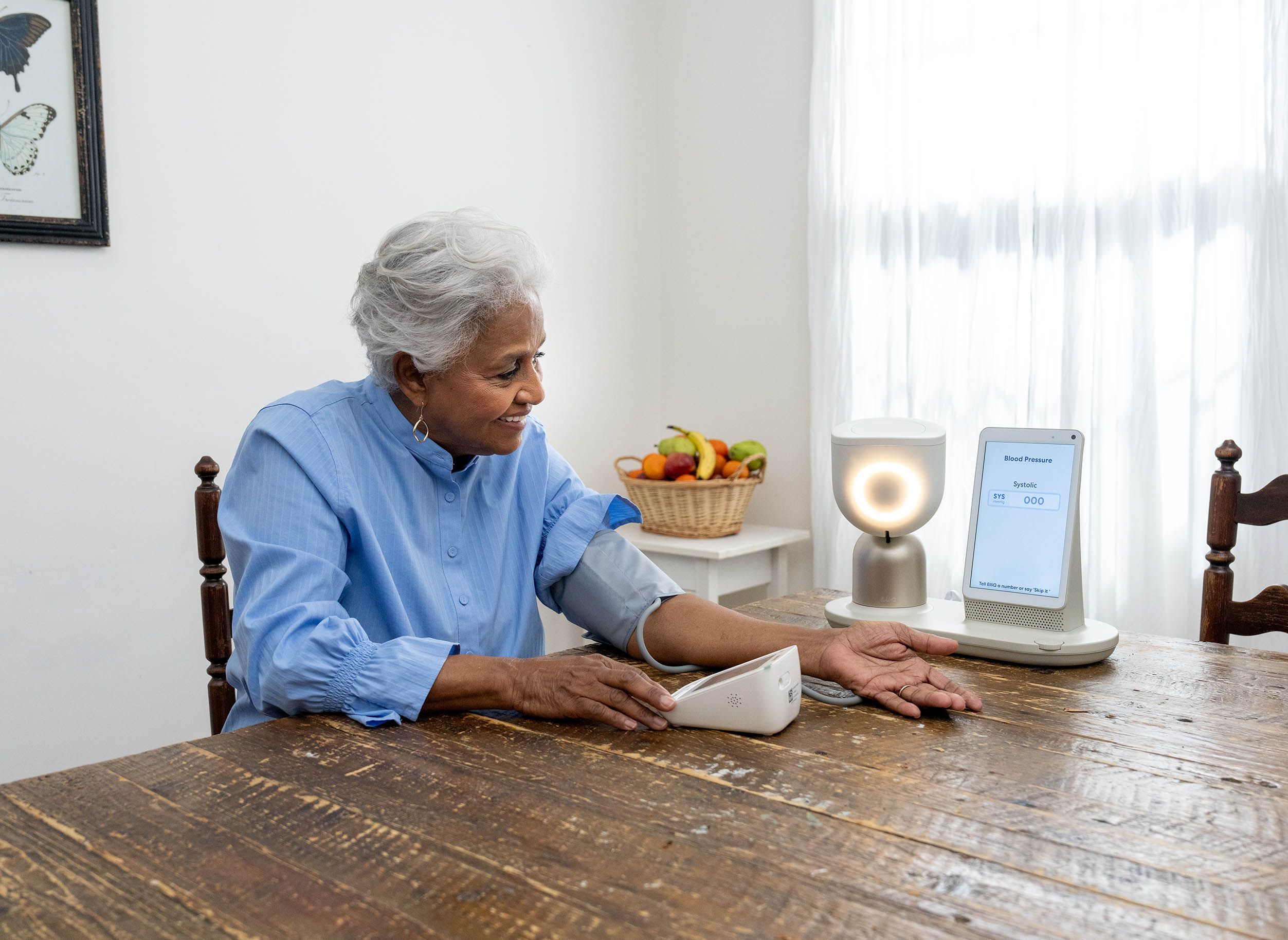 An older woman sits a table with the ElliQ system and a blood pressure cuff.
