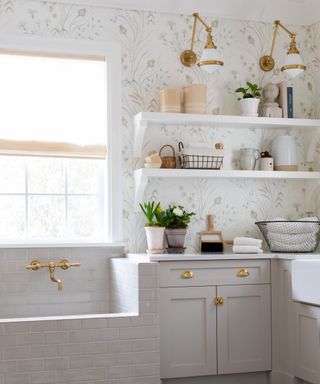 Light and breezy laundry room featuring palette of natural neutrals, subtle floral design wallpaper, and brass fittings on hardware and lighting