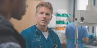 George Rainsford as Ethan Hardy in his scrubs standing in a hospital room in Casualty. 