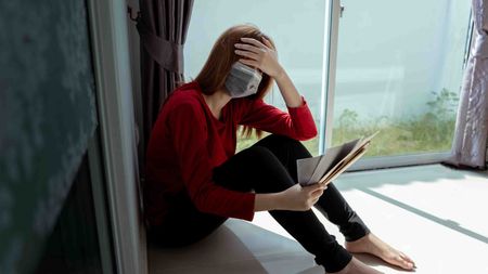 Photo of a masked woman sitting on the floor holding paperwork with her hand on her forehead