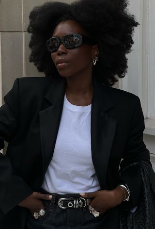 a close photo showing what to wear to the office in the summer with a woman wearing a black blazer over a white t-shirt with a hardware belt, stacked silver rings, stacked silver bangles, silver earrings, and black sunglasses