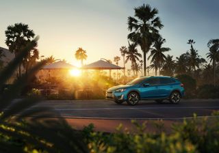 Daytime outside image of a blue XV E-Boxer Subaru plug-in hybrid at sunset, palm trees, road, parasols, shrubs, tables and chairs, blue dusk sky
