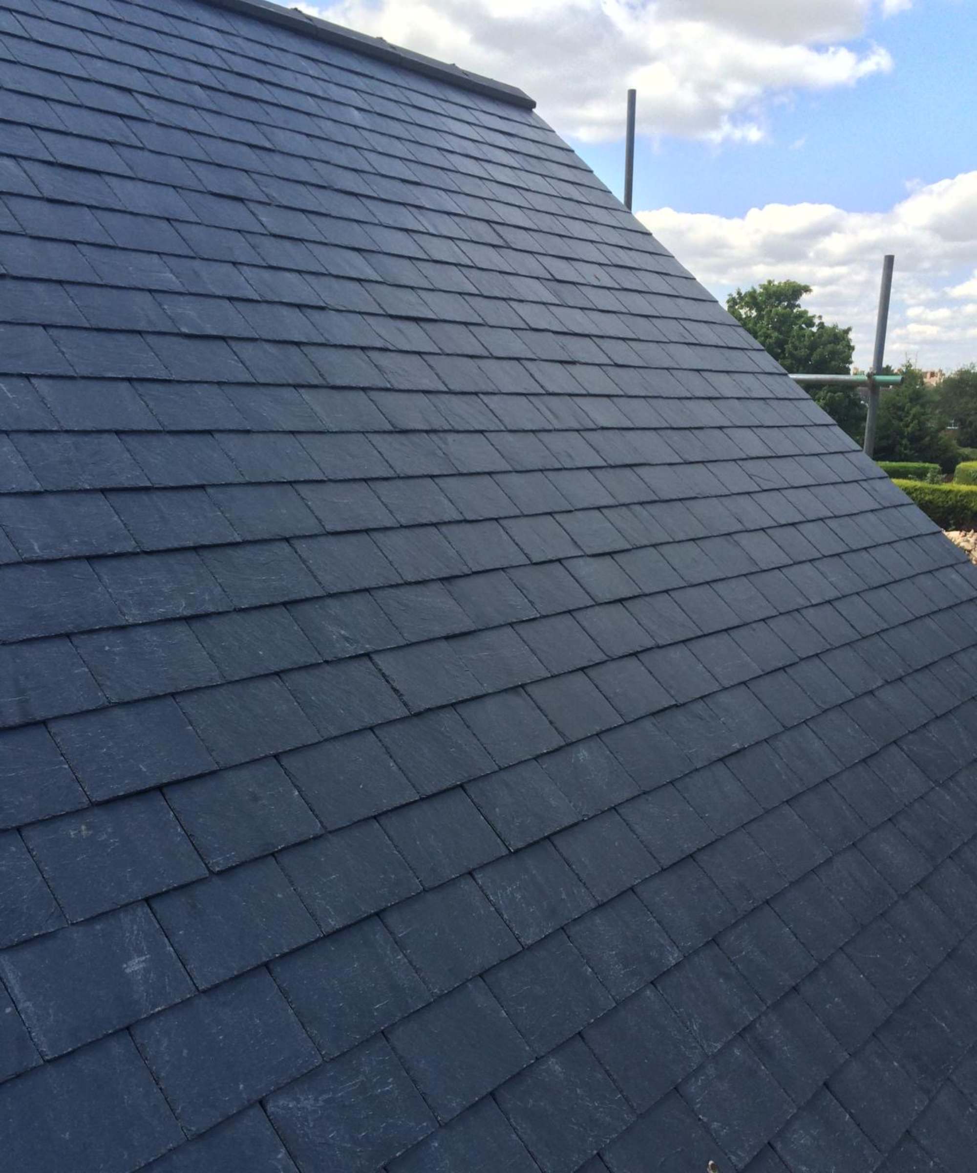 A close up of a newly slated pitched roof. The dark slate has been sourced from Northern Slate. There is scaffolding visible in the background against a blue sky.